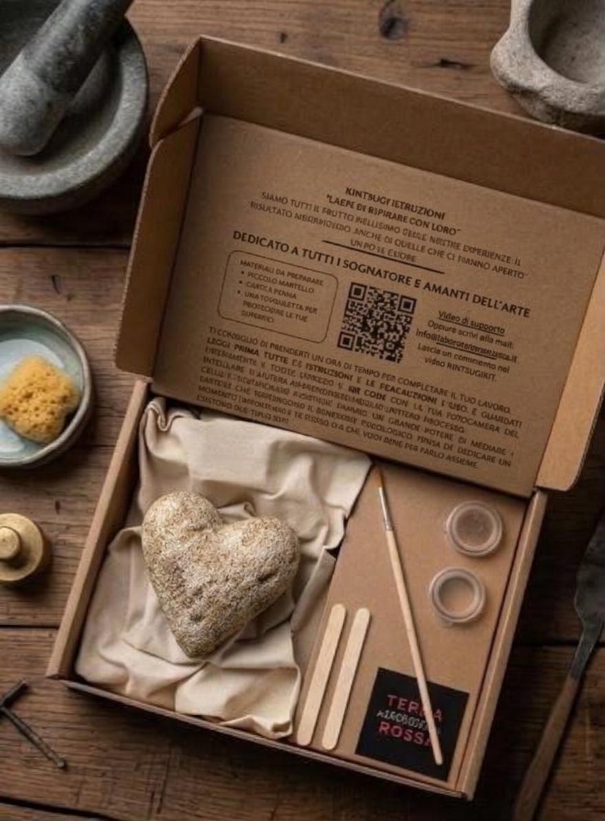 Scatola di cartone aperta con cuore di ceramica su stoffa, pennello, polvere d'oro, bastoncini e foglio di istruzioni sul coperchio